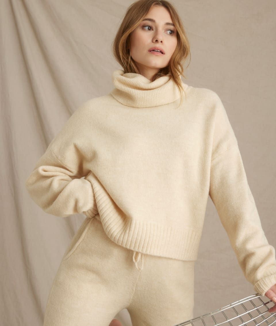 A model wears a beige turtleneck knit sweater with matching sweatpants.