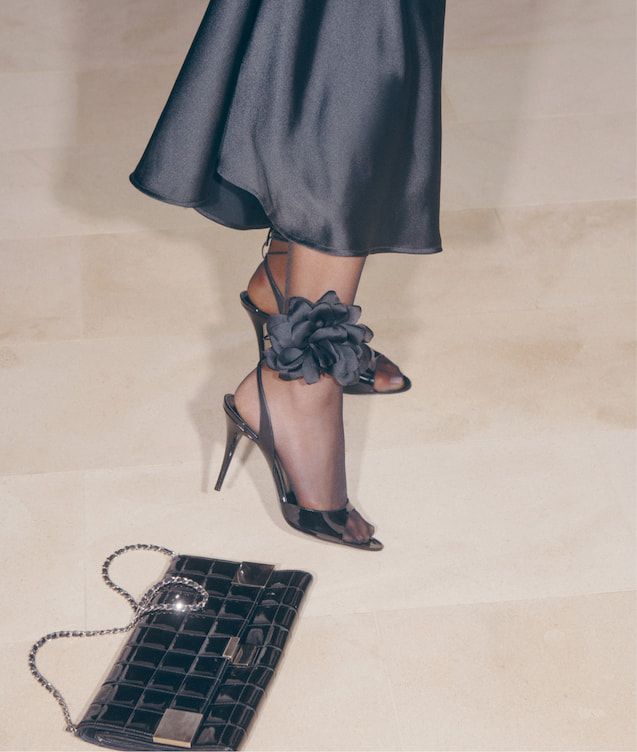An influencer wears a black satin dress with black heels detailed by a black rosette around her ankle.