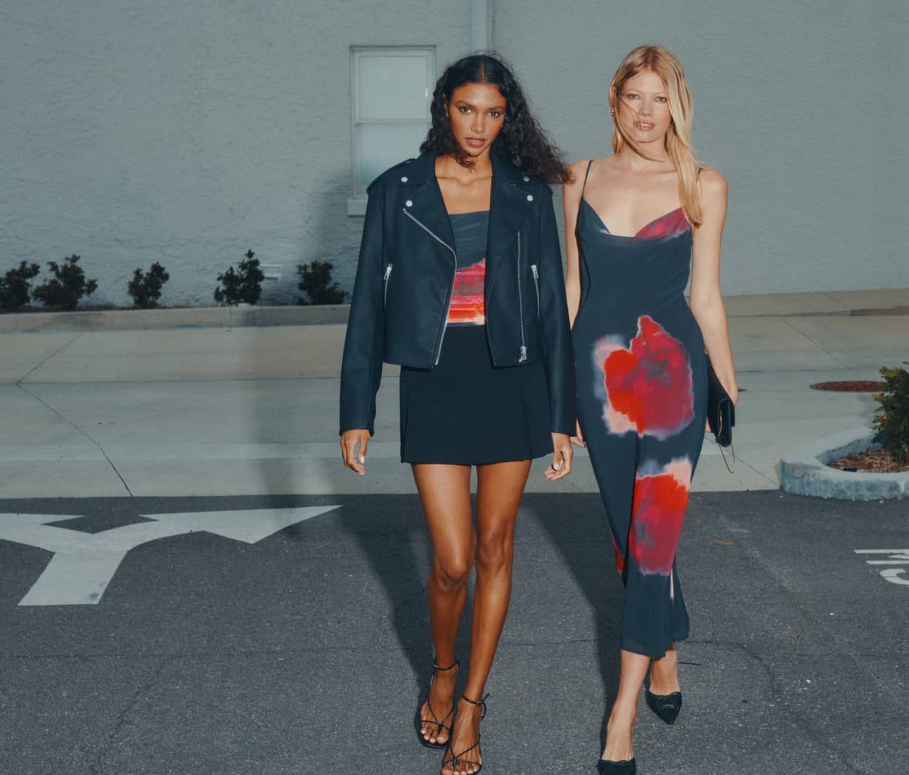 Model to the left is wearing a black faux-leather biker jacket, floral mesh top, and a black mini skort. Model to the right is wearing a black floral cowl neck maxi dress.