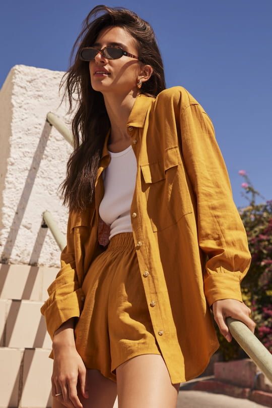 A model wears a mustard-yellow linen button-up shirt with matching linen shorts and a white tank top.