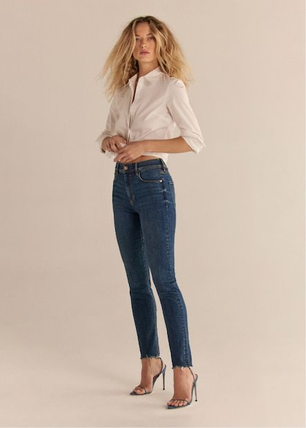 A model wears the Kate skinny jeans in dark indigo blue with a white button down shirt.