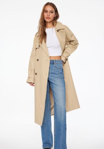 Model is wearing a beige trench coat, a white tank top, and blue jeans.