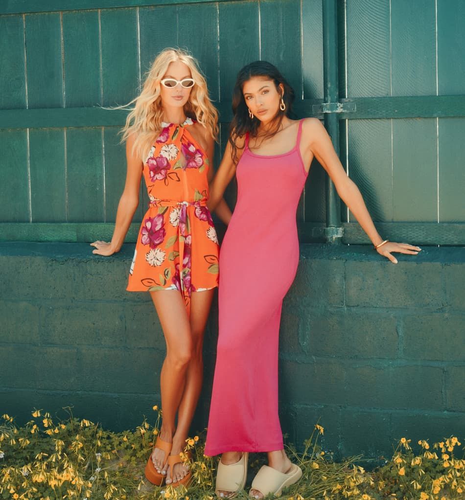 One model wears an orange floral halter neck romper and the other wears a bright pink sleeveless midi dress.