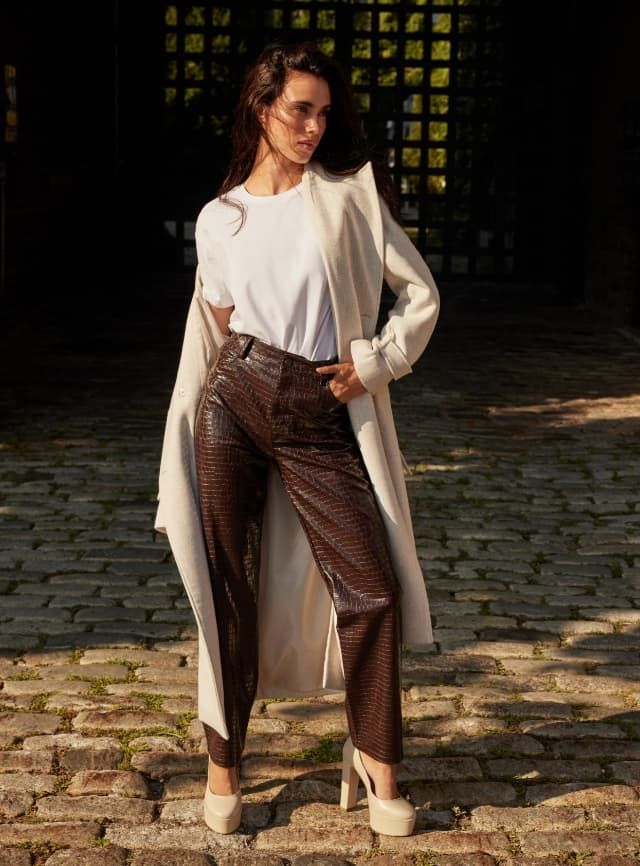 Une mannequin porte un pantalon à texture croco en faux cuir brun avec un t-shirt blanc et un manteau beige.