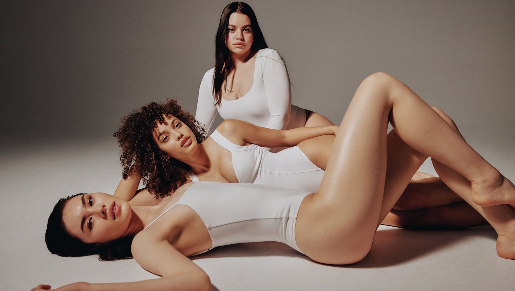 The first model wears a white cami sculpt'd bodysuit, the second model wears a white bustier sculpt'd bodysuit and the third model wears a long sleeve round neckline white sculpt'd bodysuit.