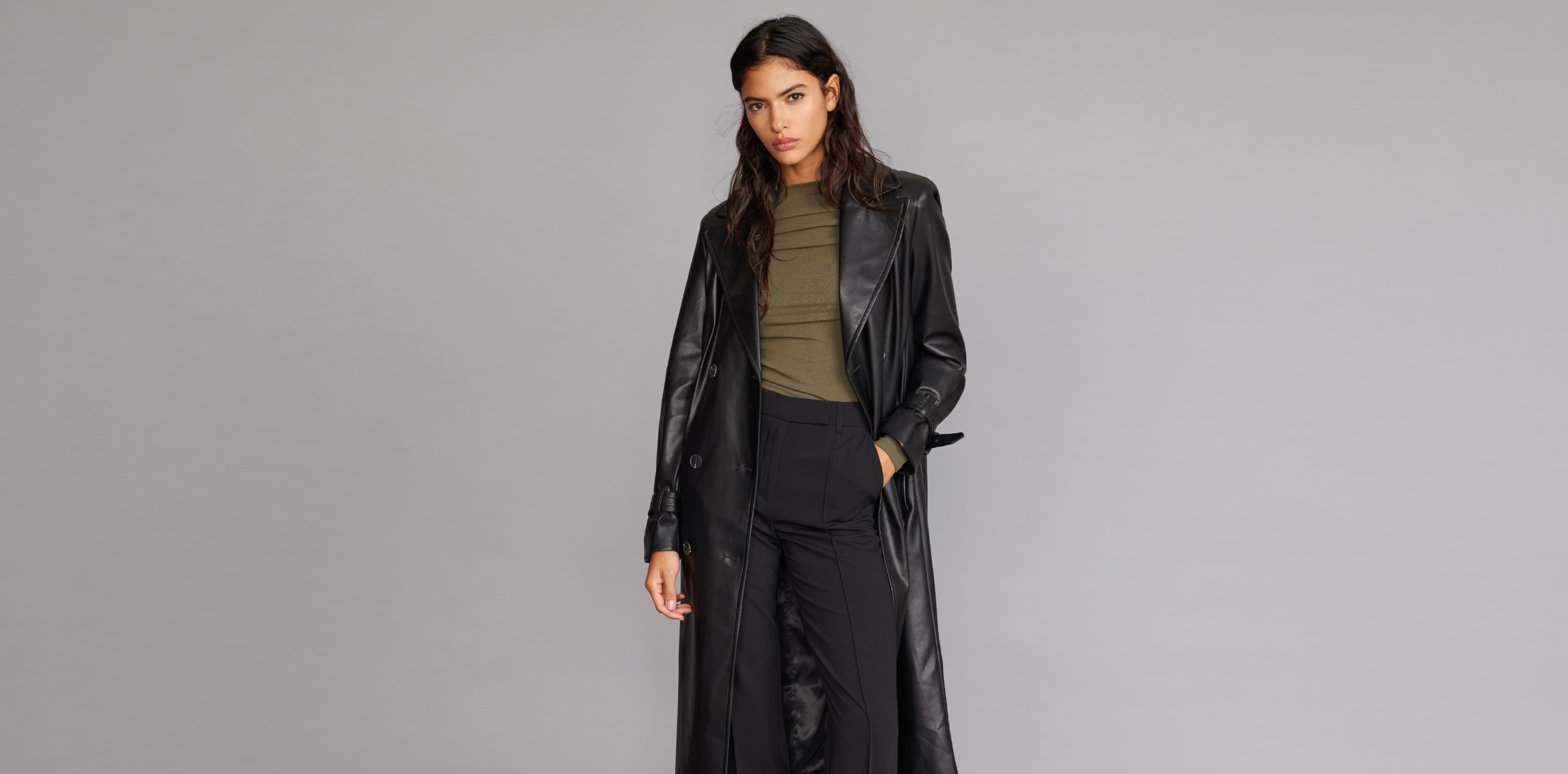 A model wears a black faux leather trench coat with black pants and a brown top.