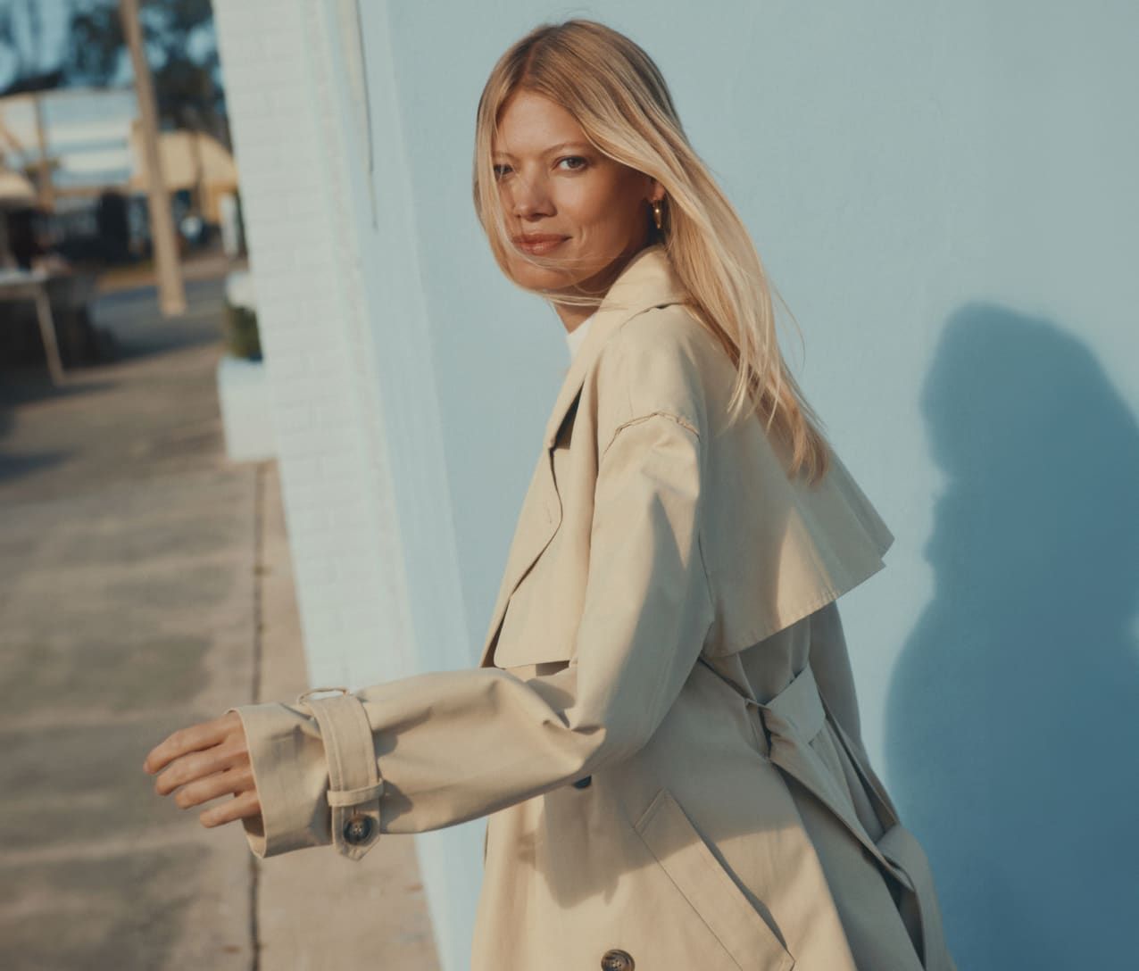 Model is wearing a beige short trench coat, a white top, and blue jeans.