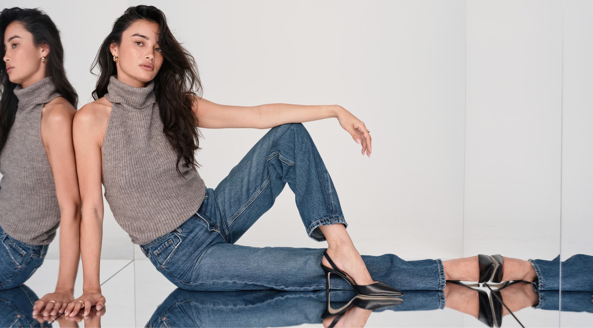 Model reclines in a sleeveless turtleneck and blue jeans.