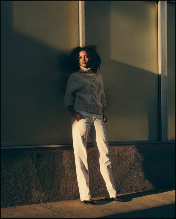 A model wears a beige turtleneck knit sweater with white wide leg jeans.