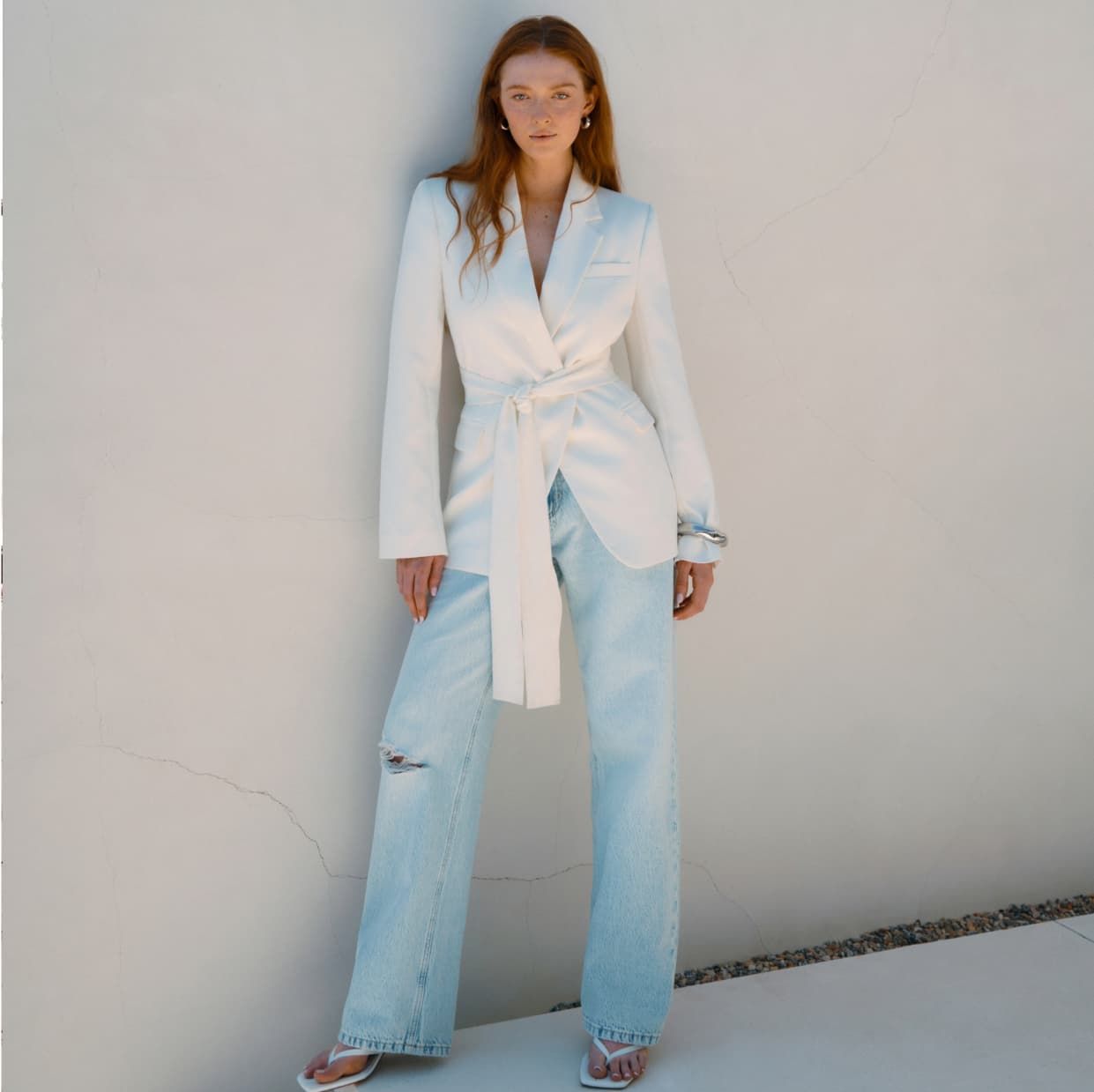 Une mannequin porte un veston blanc à ceinture avec un jean bleu pâle.