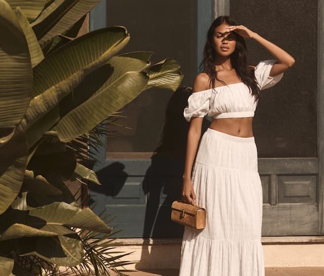 Model is wearing a matching white off shoulder crop top and tiered maxi skirt.