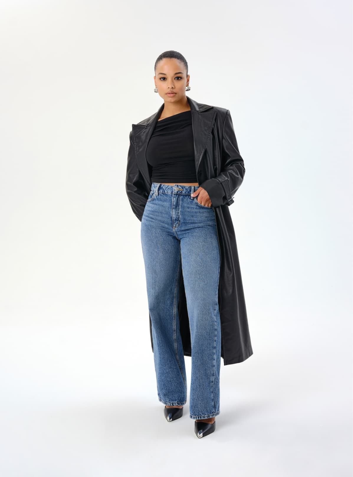 A model wears dark blue heidi jeans with a black shirt and a black long trench coat.