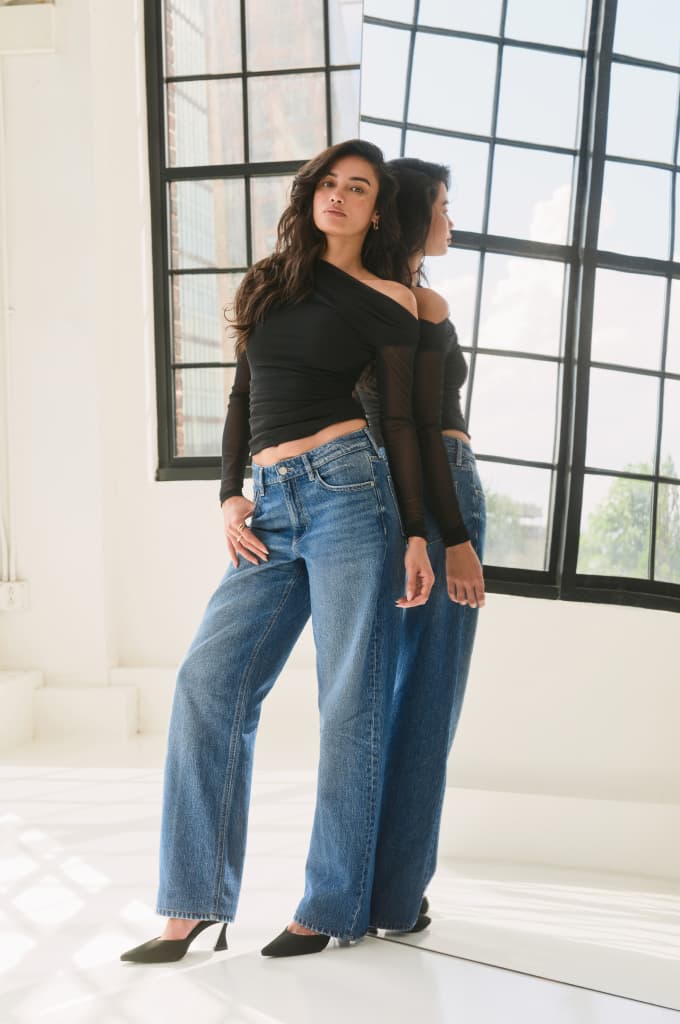 Model is wearing a black off shoulder mesh top and blue relaxed straight leg jeans.