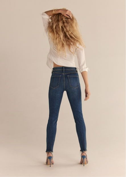 A model wears the Kate skinny jeans in dark indigo blue with a white button down shirt.