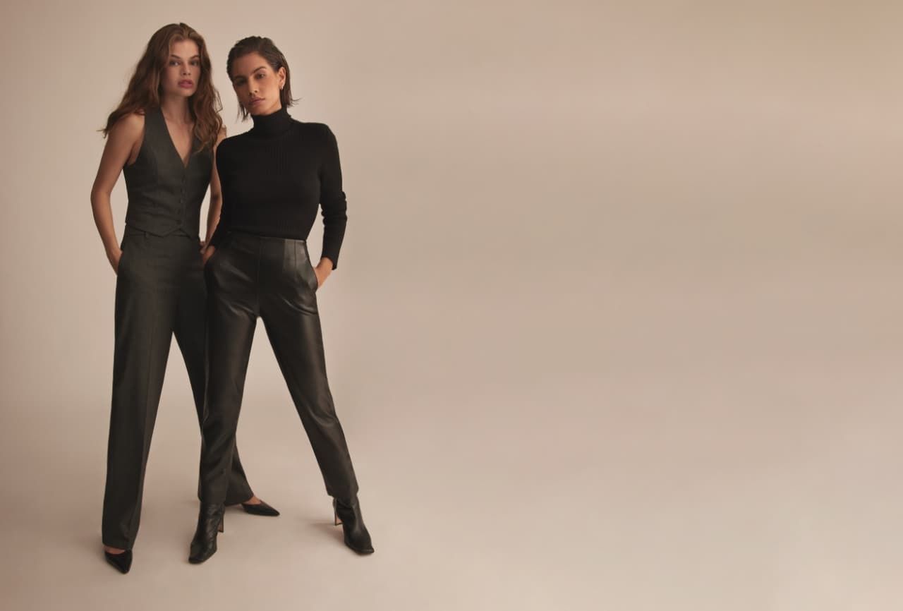 One model wears grey slim leg pants with a macthing grey vest and the other model wears faux leather black pants with a black turtle neck.