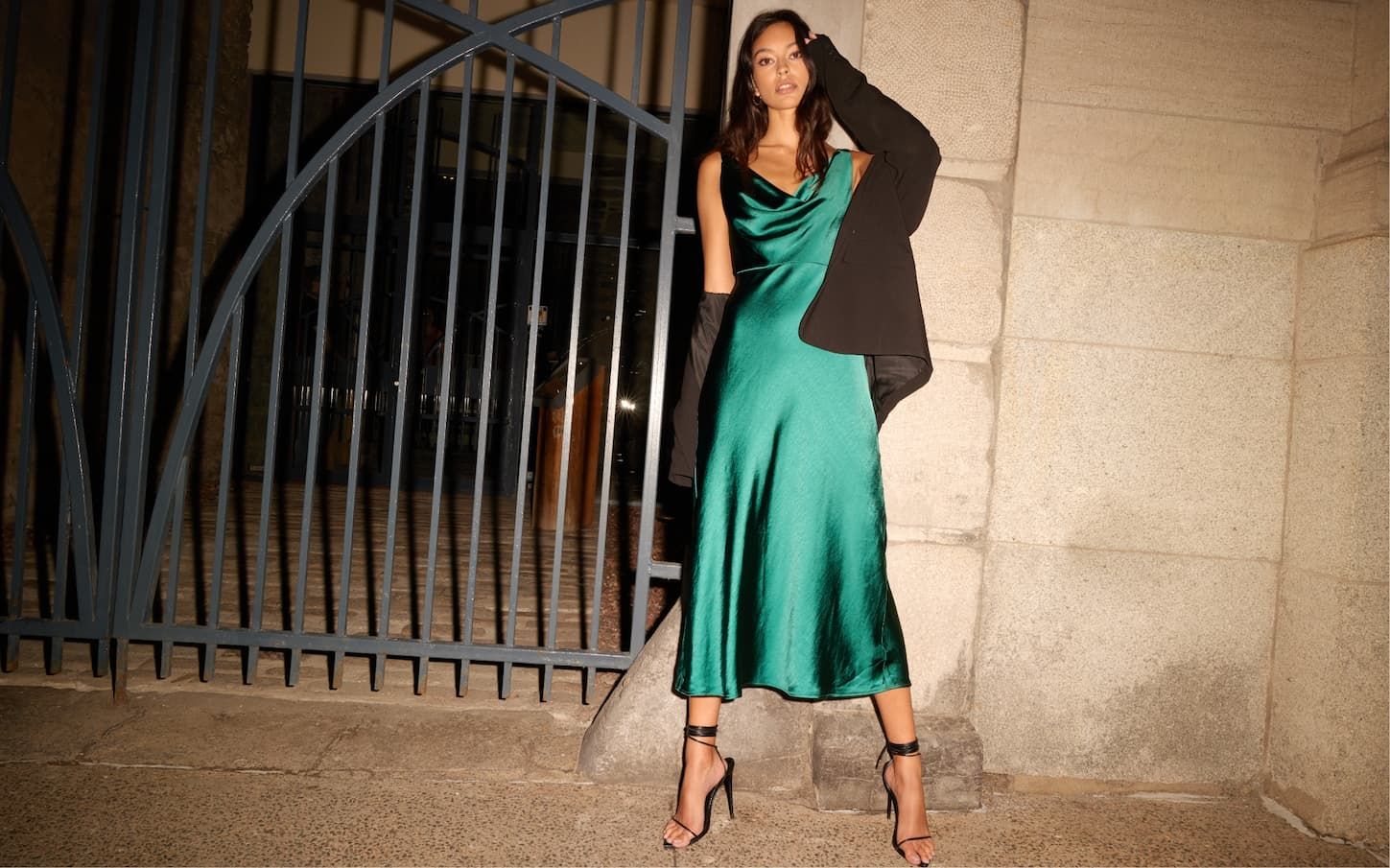 A model wears a long green satin cowl neck dress with a black blazer over.