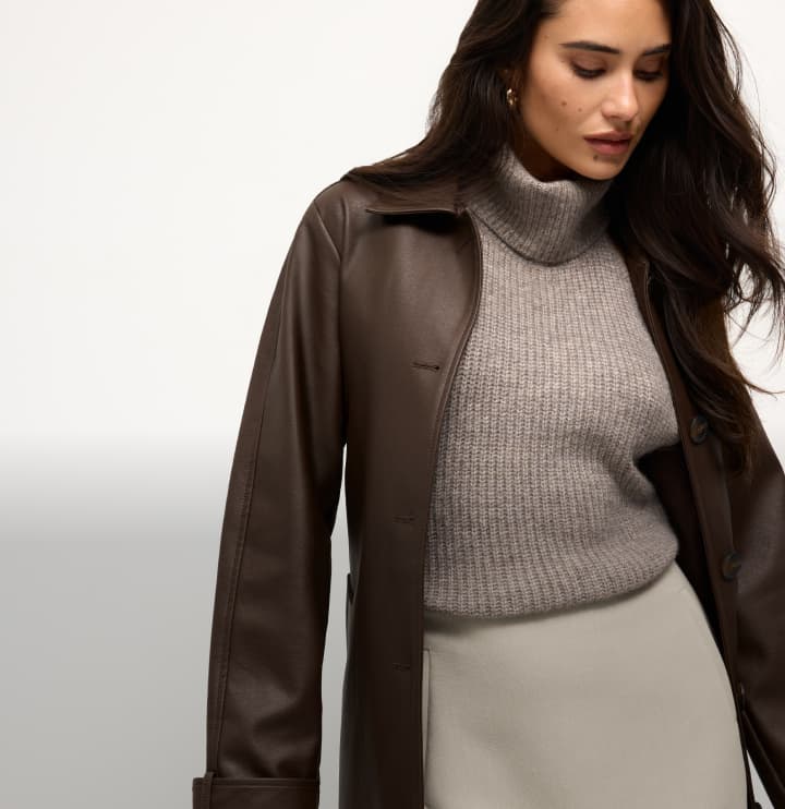 Model is wearing a brown faux leather coat over a brown turtleneck and white mini skirt.