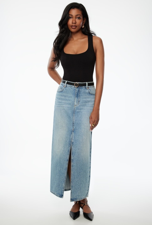 A brunette woman in a black dress and heels stands posing.