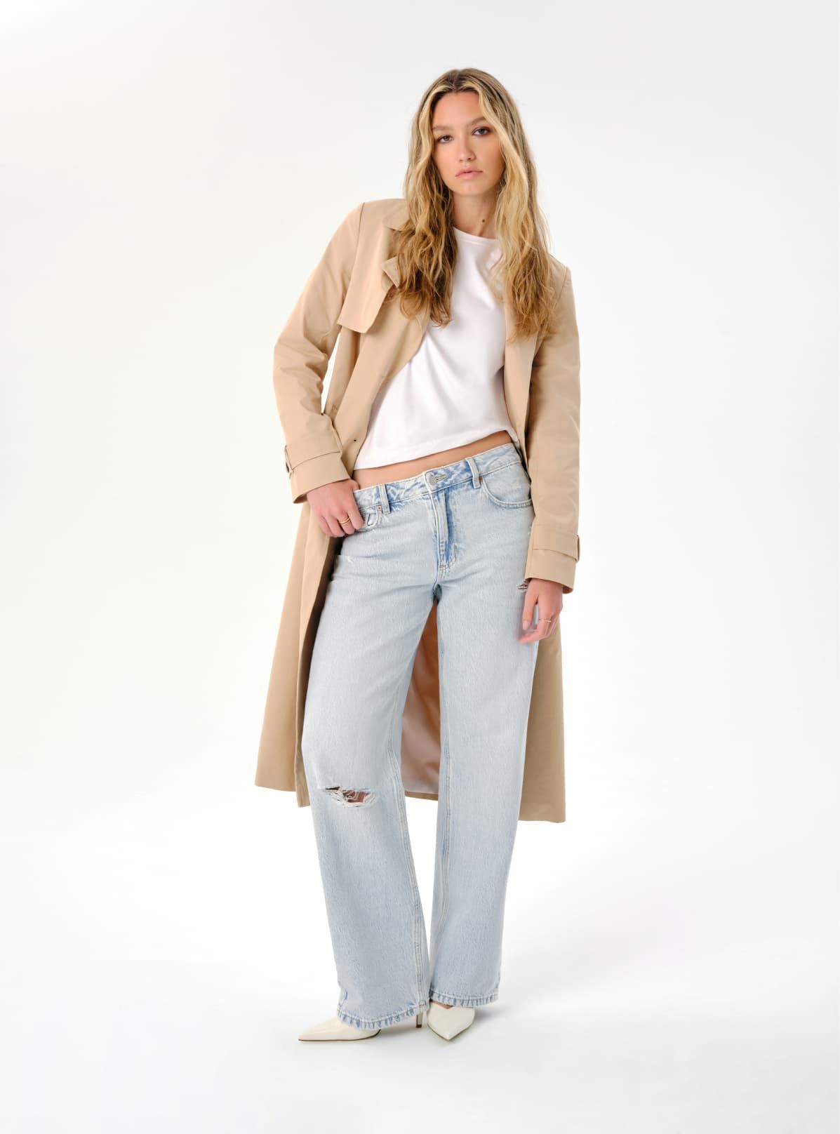 A model wears light blue distressed relaxed jeans with a white shirt and beige trench coat.