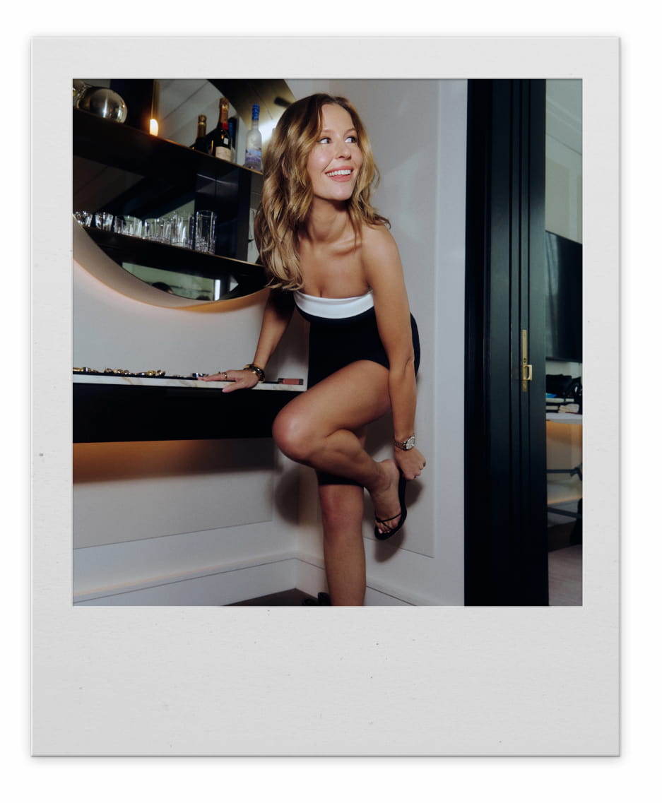 A blonde woman smiles while lifting her foot to adjust her sandal heel.