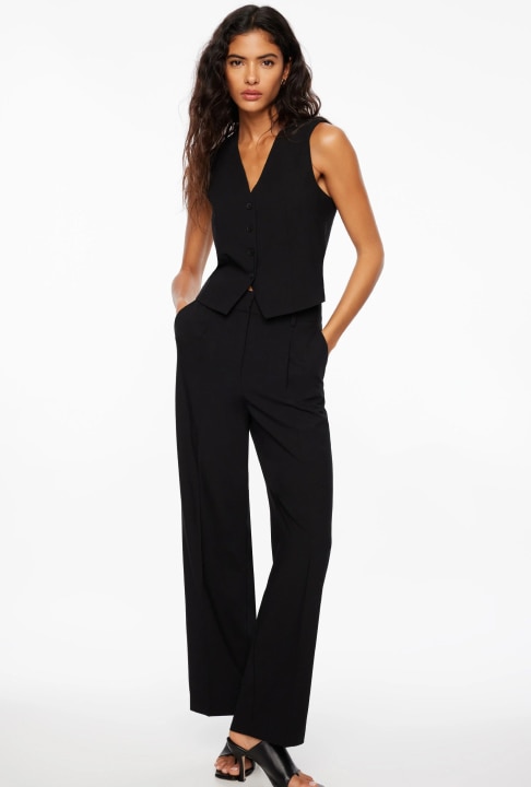 A woman in a black vest and pants stands posed with her hands in her pockets.