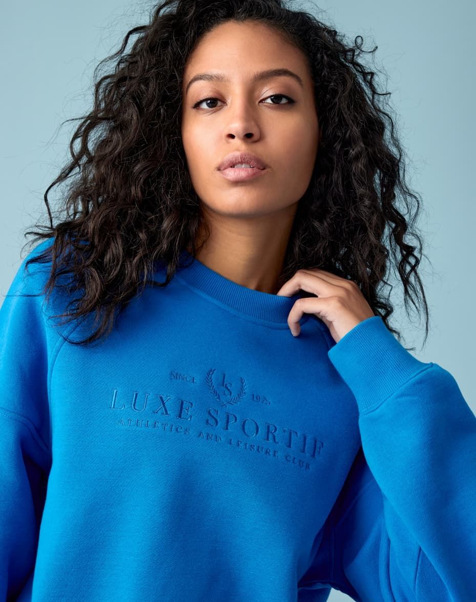 A model wears a blue crewneck with 'athletics & leisure club' embroidered.