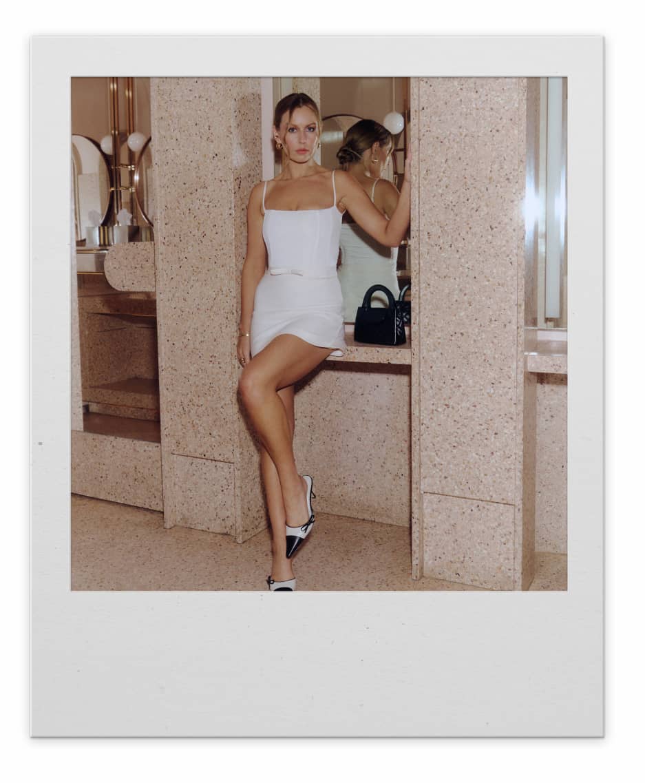 A brunette woman in a white mini dress poses leaning against a column. 