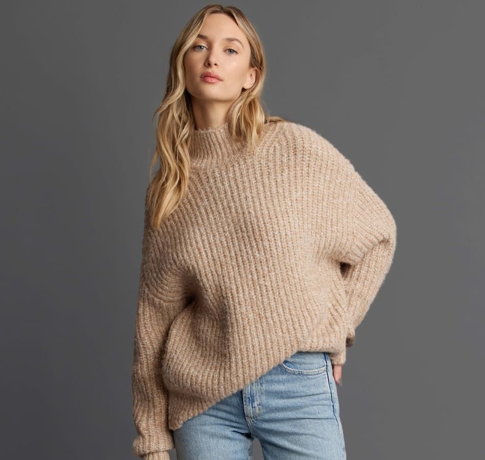 A model wears a beige mockneck sweater with blue jeans.